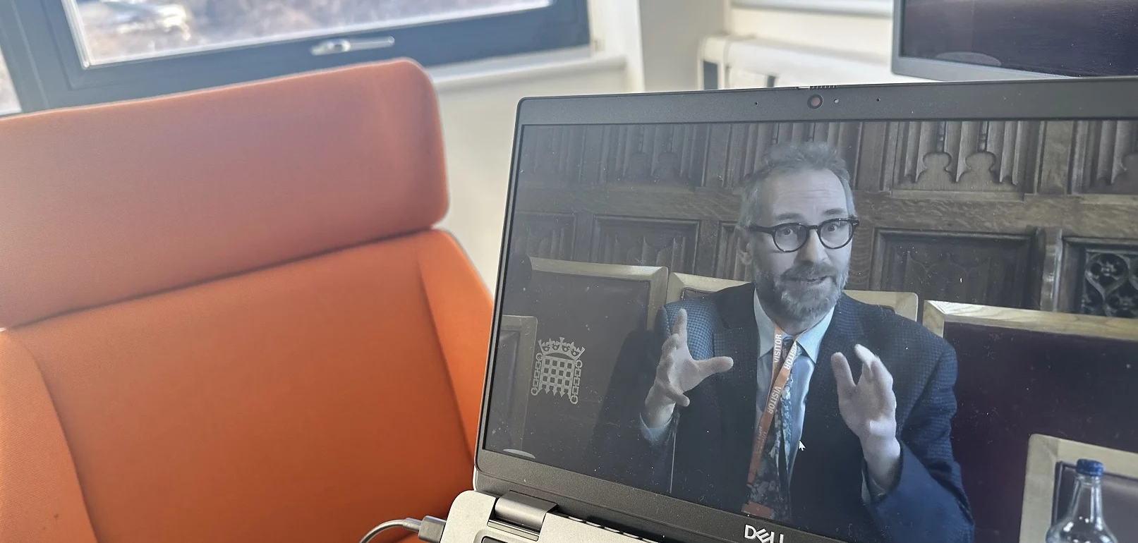 A laptop screen displaying SEI’s Kevin Hicks giving evidence at the UK House of Lords nitrogen inquiry. He is seated in a formal setting, gesturing with both hands while speaking. In the background, an orange chair and a window are visible in a well-lit office space.