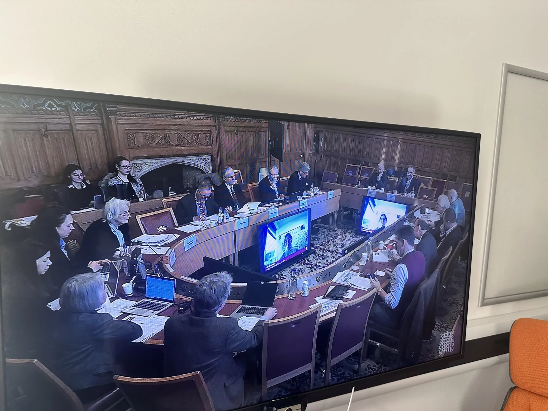 A screen displays a formal parliamentary session in the UK House of Lords, where experts and officials are gathered around a large, curved table. Several participants are taking notes, using laptops, and listening to the discussion.