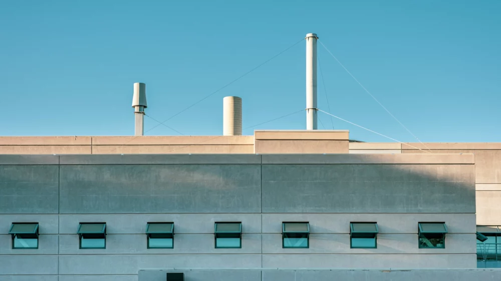 The top part of a grey building with seven open windows, all the same size. Three silver ventilation shafts of different sizes extend upwards from the roof into a blue sky.