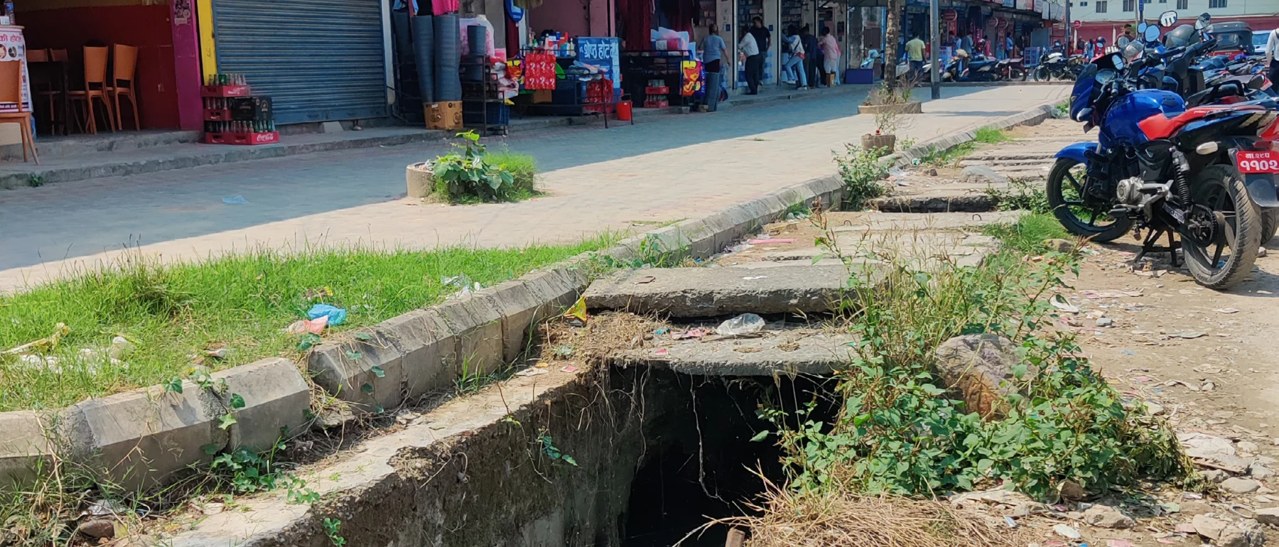 An open canal with water runs between a sidewalk lined with colorful storefronts and the street, with motorbikes parked nearby.