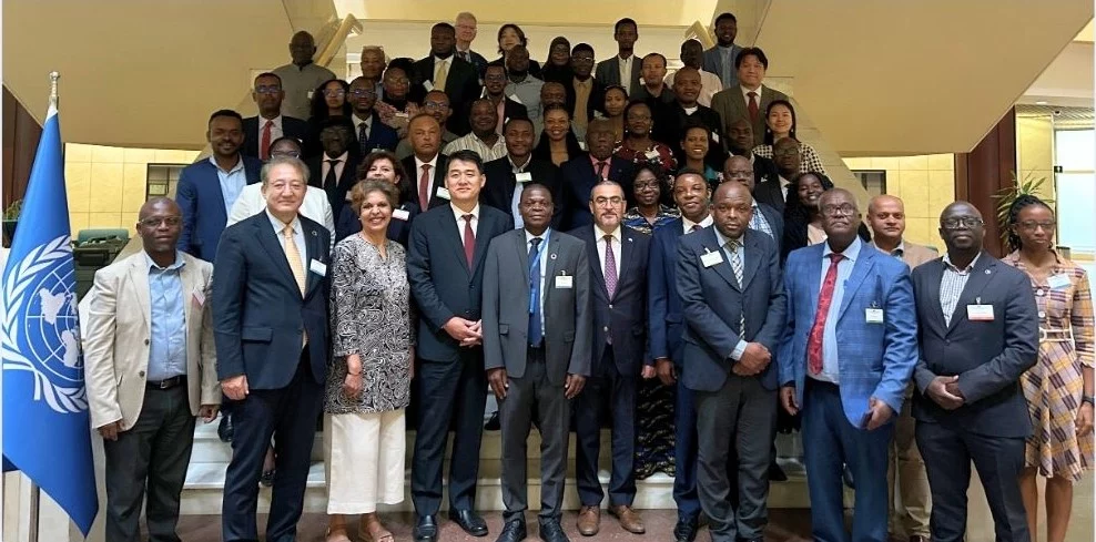 A large group of people standing on a staircase posing for a formal group photo at a UN event. A UN flag is visible on the left side of the image.