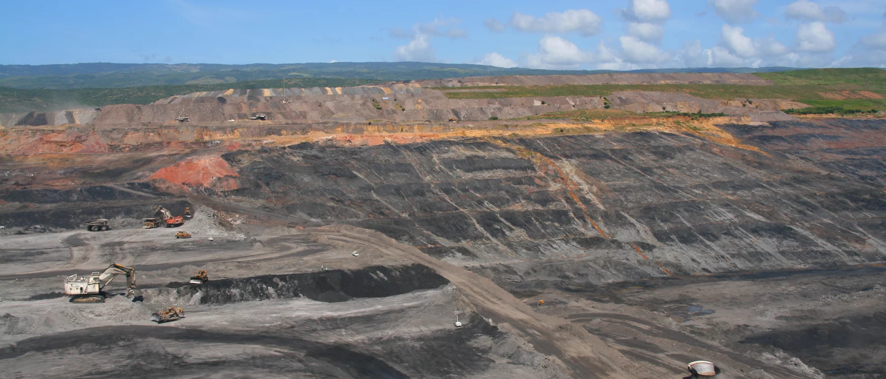 A large open-pit coal mine with terraced slopes, mining equipment, and a dirt road under a blue sky.