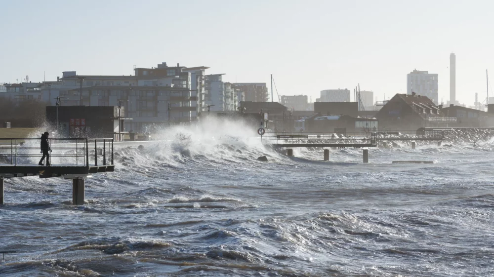 Stormy waves crash against a pier in a coastal urban area with a lone person walking on the platform.