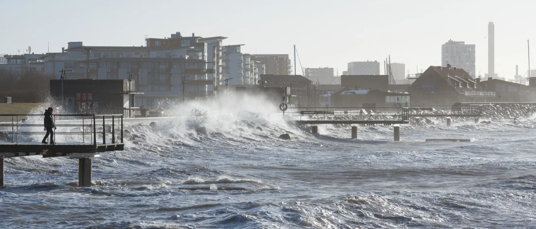 Stormy waves crash against a pier in a coastal urban area with a lone person walking on the platform.