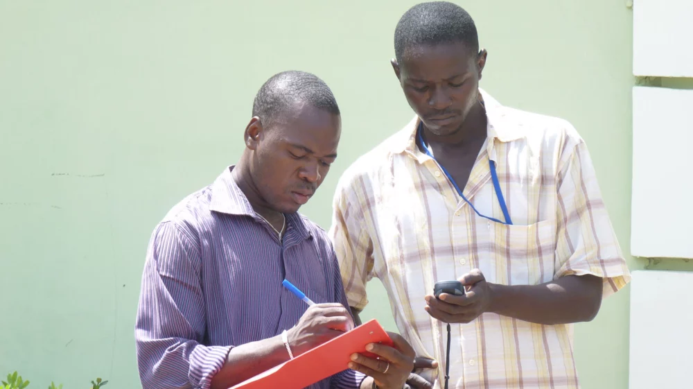 Two men stand together outdoors. One is making notes on a red clipboard, the other is reading out information to him from a small data-collection device.