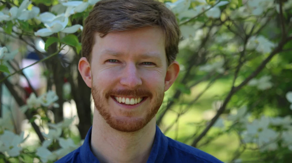 Portrait of Taylor, smiling at the camera in front of a tree, wearing a royal blue button-up shirt.