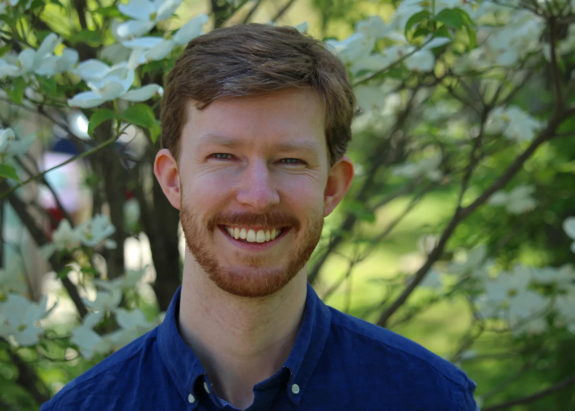 Portrait of Taylor, smiling at the camera in front of a tree, wearing a royal blue button-up shirt.