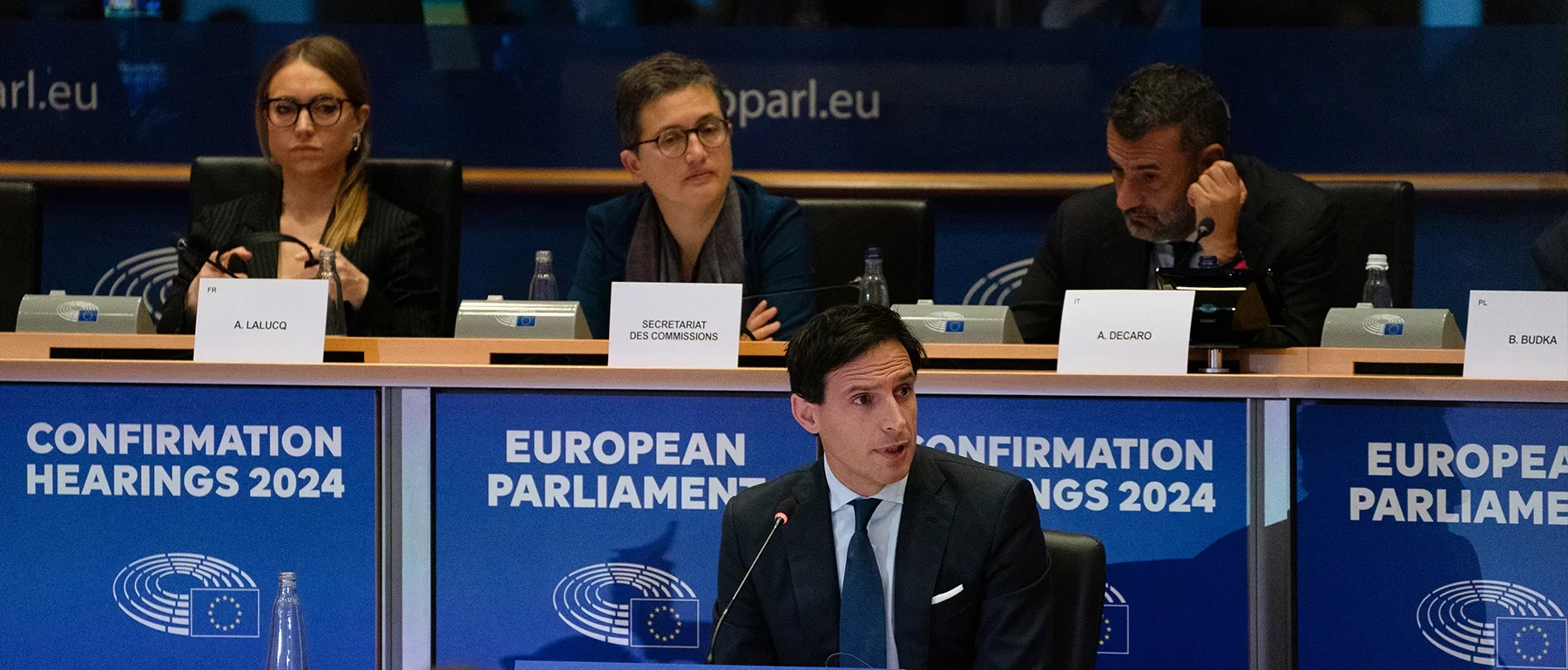 Wopke Hoekstra speaking at the European Parliament’s 2024 confirmation hearings, seated at a panel with other officials and blue signage behind.