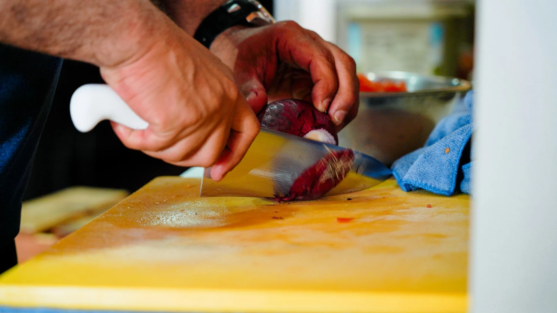 close up of a man's hands using a large knife to cut a red onion on a wooden chopping board.
