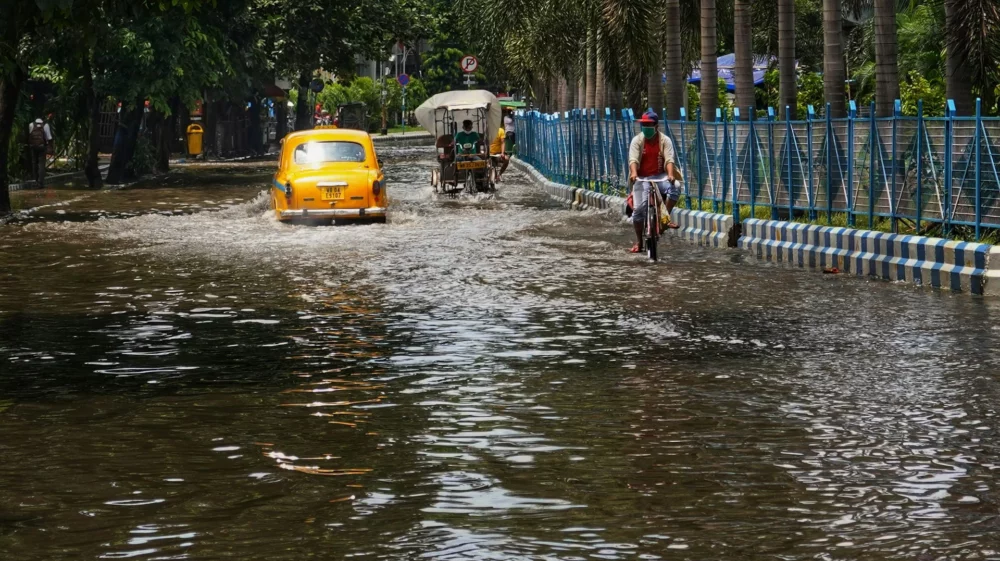 People navigate a flooded street in an urban area, with a yellow taxi, a cycle rickshaw, and a man riding a bicycle through the water. Trees and a fence line the road.