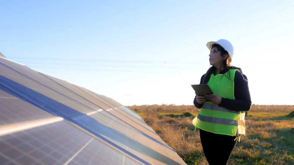 Woman in reflective yellow vest and white hard hat examines a solar panel in a grassy field