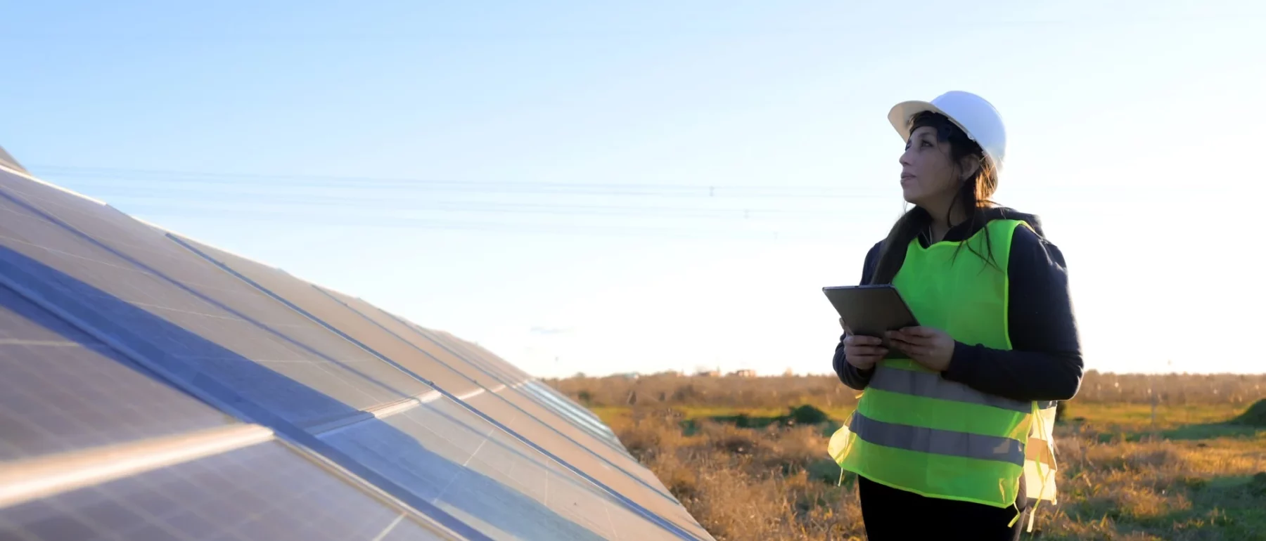 Woman in reflective yellow vest and white hard hat examines a solar panel in a grassy field