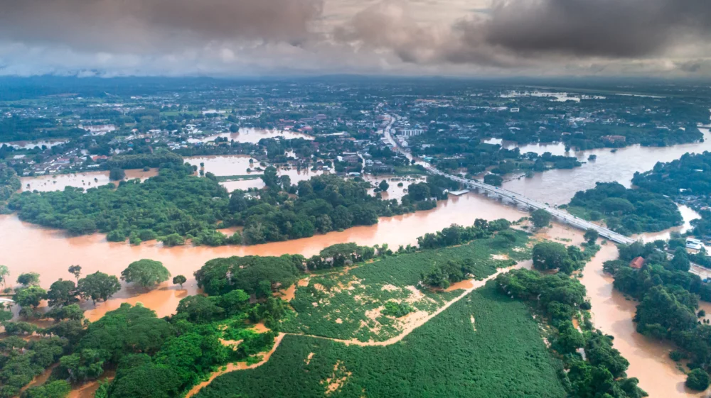 Aerial view of a flooded landscape with muddy rivers overflowing into nearby green fields and urban areas under cloudy skies.