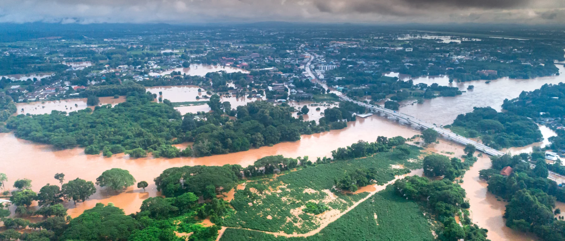 Aerial view of a flooded landscape with muddy rivers overflowing into nearby green fields and urban areas under cloudy skies.
