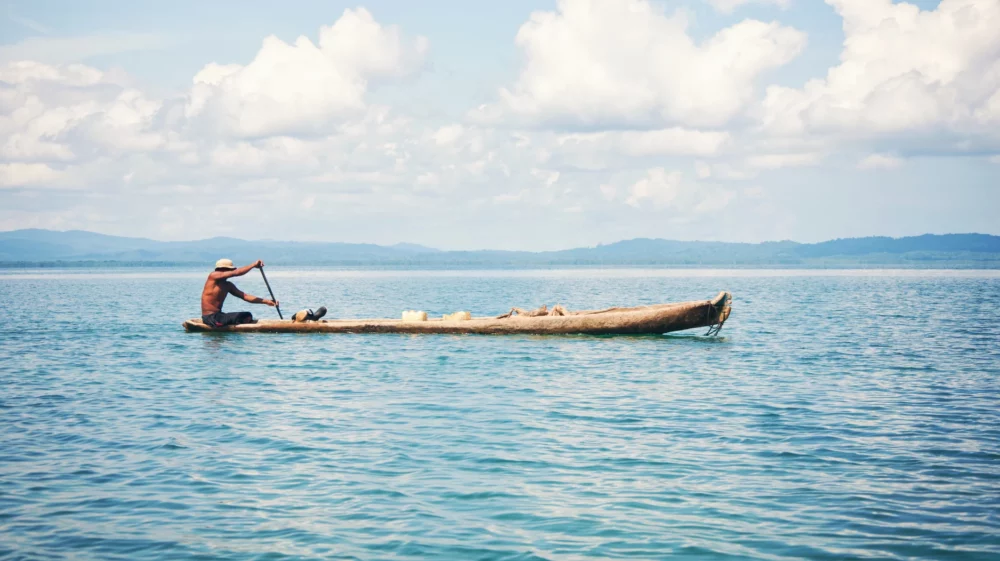 A man paddles a traditional wooden canoe across calm blue waters, with distant forested hills under a partly cloudy sky.