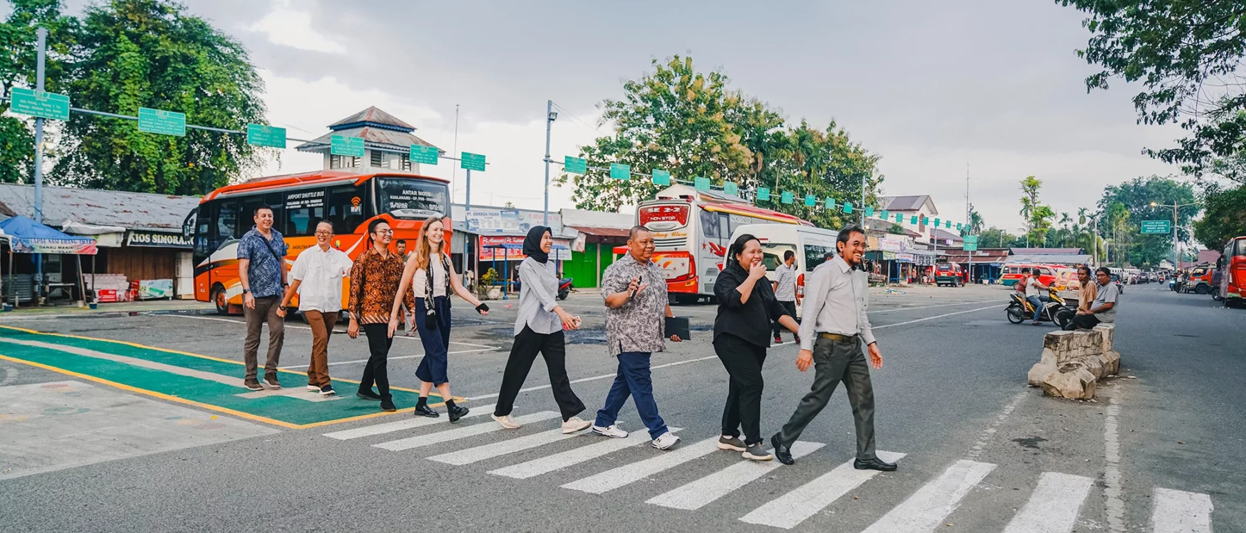 Group of people smiling as they walk across a zebra crossing near buses and market stalls in a public transport terminal in Medan, Indonesia.