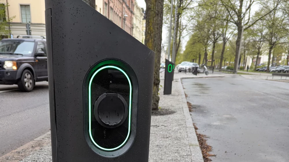 Close-up of a modern electric vehicle charging station with a glowing green light, located on a city street lined with trees and parked cars on a rainy day.