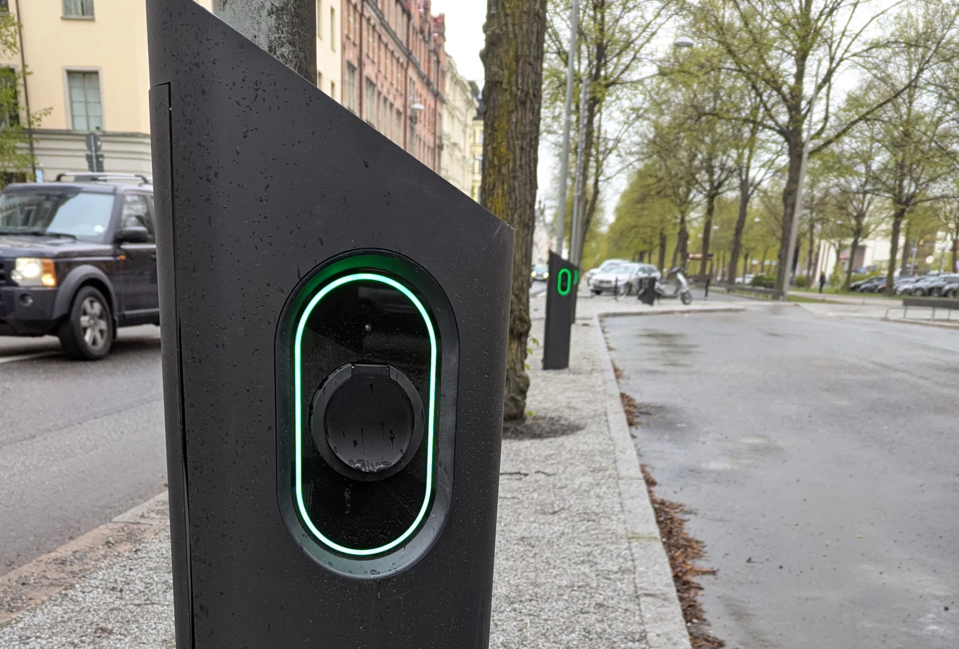 Close-up of a modern electric vehicle charging station with a glowing green light, located on a city street lined with trees and parked cars on a rainy day.