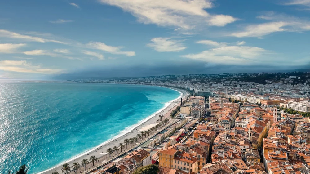 A scenic coastal city with turquoise-blue sea, a palm-lined beach promenade, and densely packed buildings with red-tiled roofs, under a partly cloudy sky.