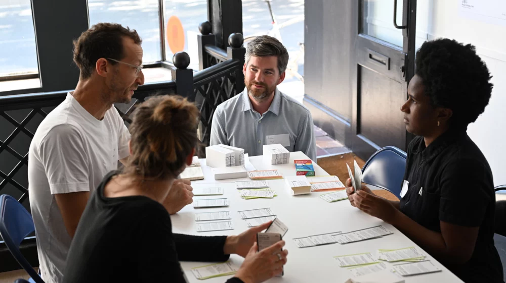 Three people (two women and one man) sit around a table while in discussion. Another man is crouching at the head of the table, listening to and facilitating the discussion.