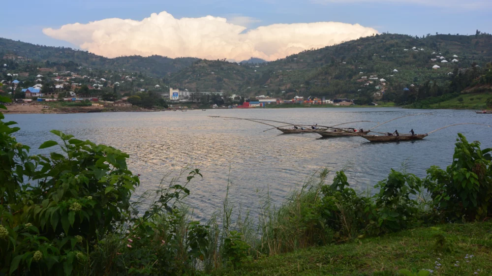 Three-hulled fishing boat leaves its harbor at sunset on Lake Kivu, Rwanda, under a blue sky with white clouds, against lush green hills dotted with buildings.