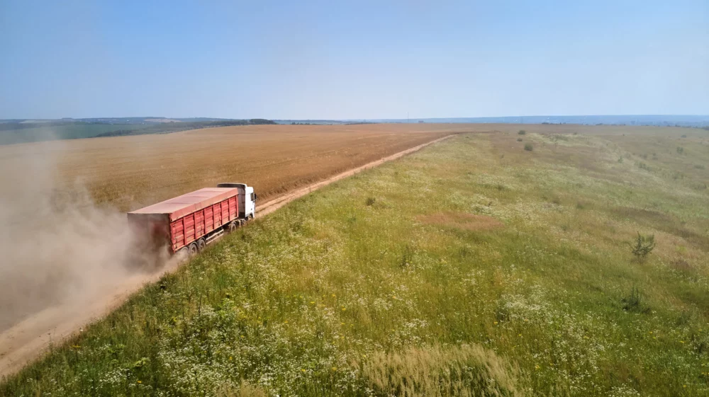 Aerial view of lorry cargo truck driving on dirt road between agricultural wheat fields, green to the right and brown to the left, with the slight curve of a city on the horizon.