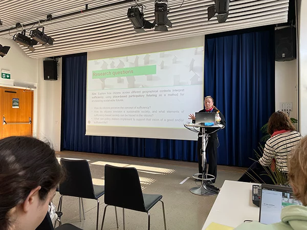 Julia Jokiaho standing at a podium presenting a slide with research questions on participatory futuring and sufficiency in a conference room.