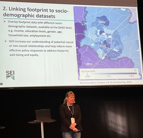 Katarina Axelsson speaking in front of a projected slide showing a map of Lund and text about linking carbon footprints to socio-demographic data.