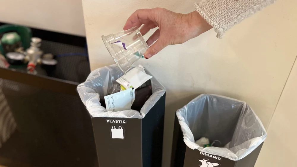 A hand holding a plastic cup hovers above a black recycling container labeled plastic, filled with trash, beside another labelled organic.