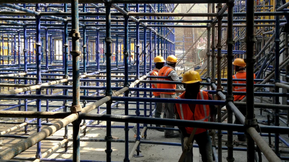 Construction workers in safety gear navigating through a dense grid of steel scaffolding at a building site.