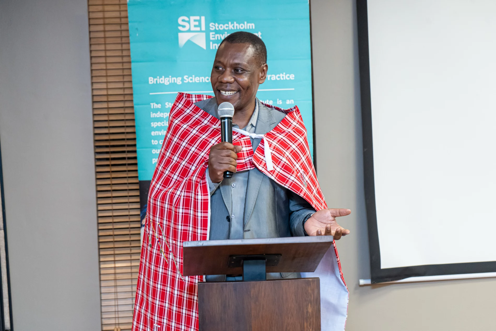 A man wearing a red and white checkered shawl speaks at a podium with a microphone, standing in front of an SEI banner.