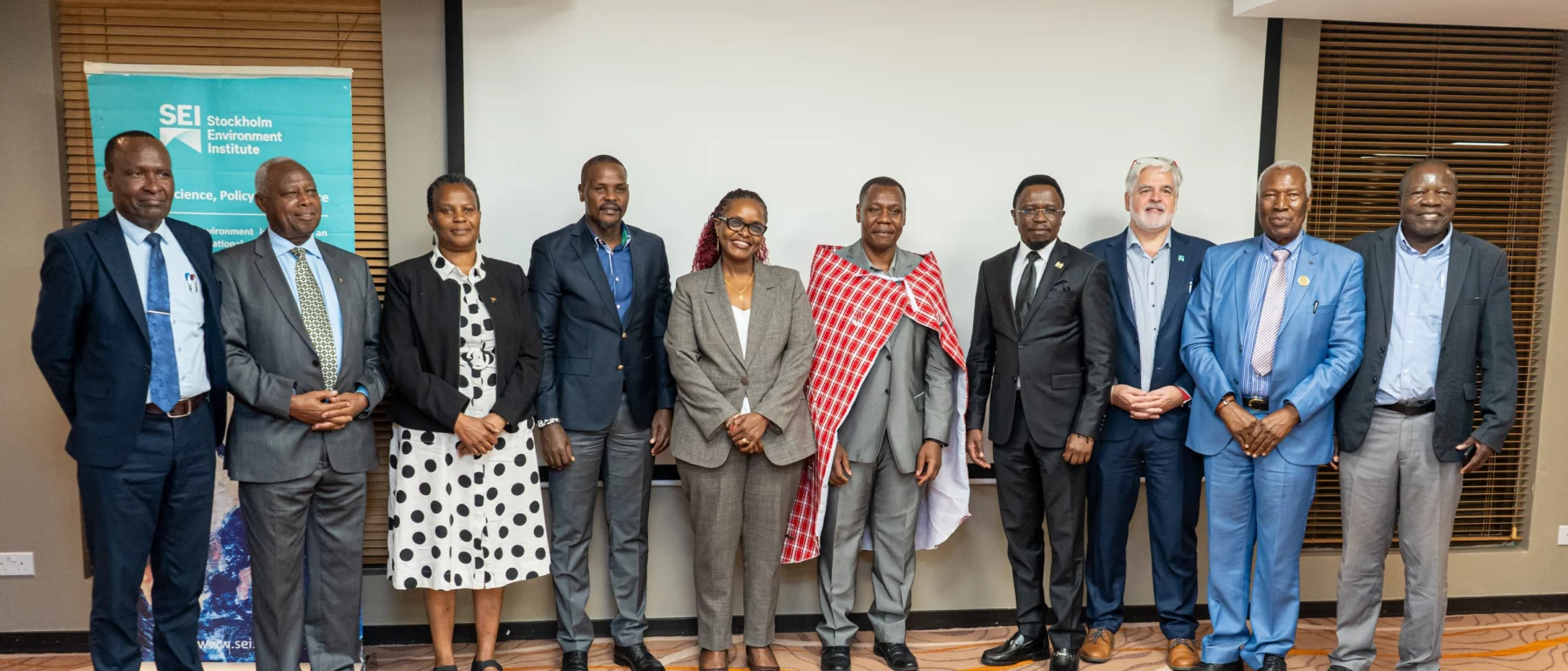 A group of eleven professionally dressed individuals stands in a line for a group photo at an event. One person, wearing a red and white checkered traditional shawl, is standing in front of a backdrop with the SEI logo.