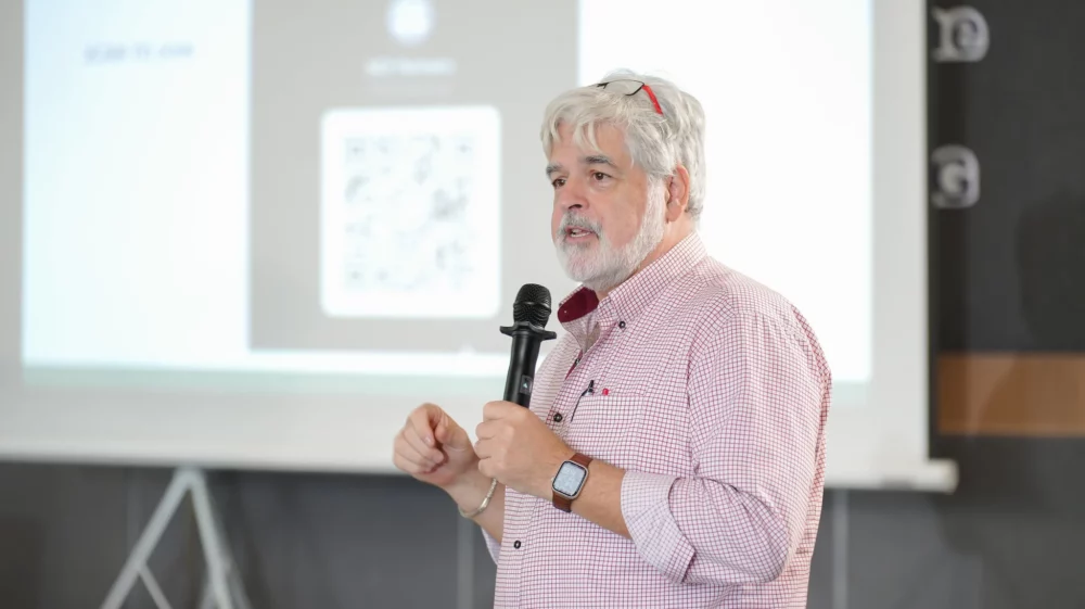 Man speaking into a microphone during a presentation, with a QR code projected on the screen behind him.