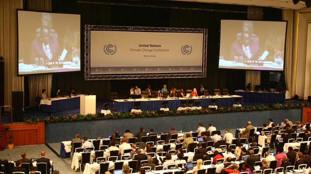 A large conference hall filled with delegates seated at rows of tables during a plenary session of the UN Climate Change Conference in Bonn, May 2012. A large screen displays a speaker, and the backdrop features the UN Climate Change logo.