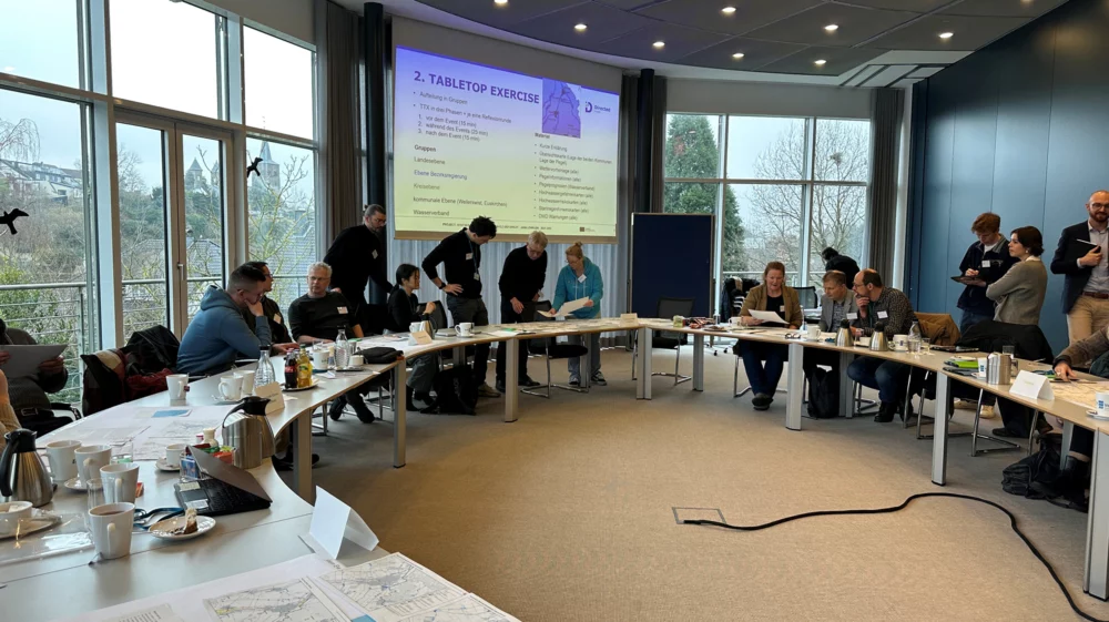 People gathered in a conference room participating in a tabletop exercise, with documents, maps, and coffee on tables arranged in a U-shape; a presentation slide is projected at the front of the room.
