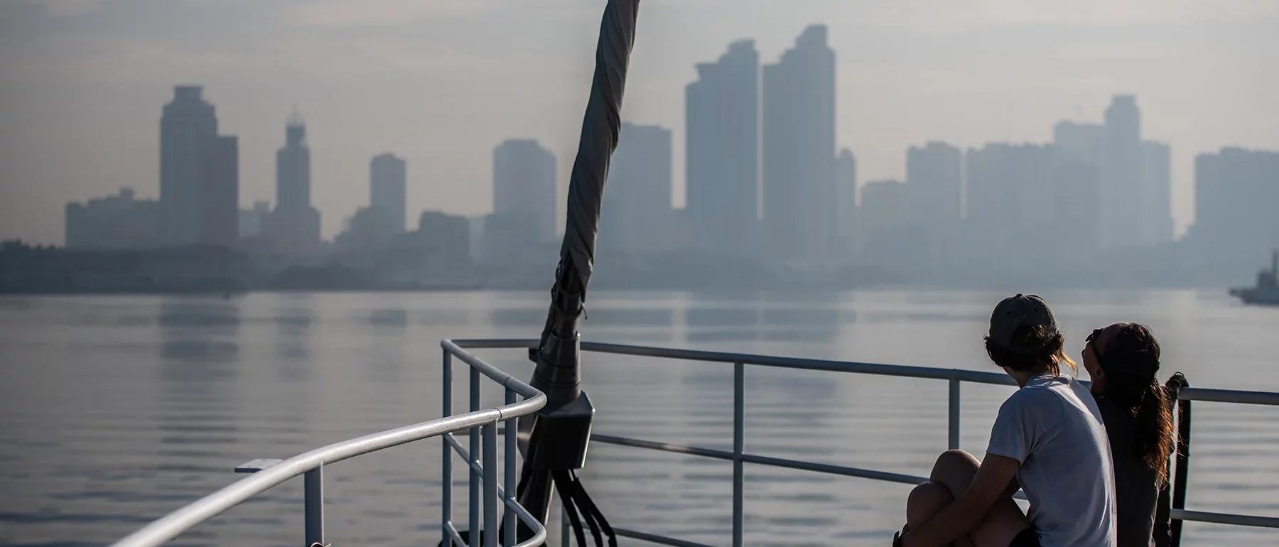 Two people sitting on the deck of a boat, looking towards a hazy city skyline across calm water.