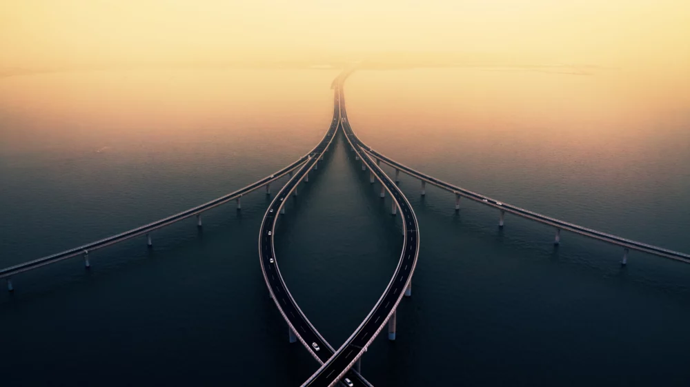 Aerial view of an elegant, multi-lane bridge with curved ramps converging and extending across calm water under a hazy, golden sky.