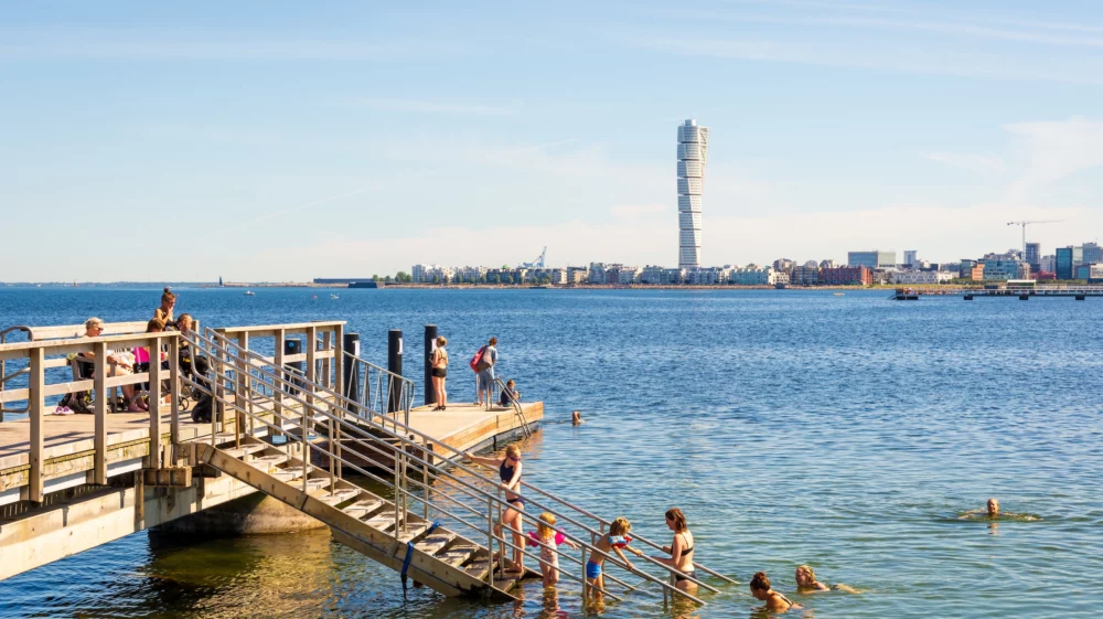 Malmö, Sweden - July 20, 2024: Adults, children and seniors enjoy sea bathing on a sunny summer day at the pier 6 of the Ribersborg beach, with a view over the Turning Torso tower.
