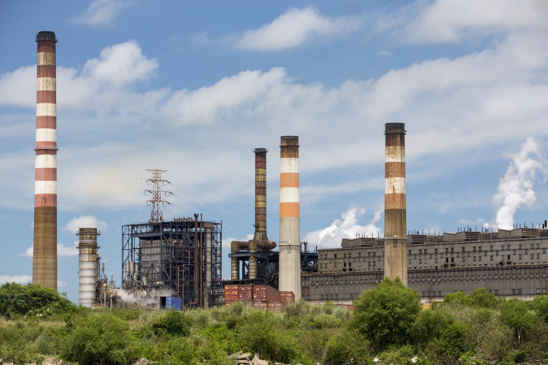 An industrial facility with multiple smokestacks emitting white smoke, set against a blue sky with scattered clouds and greenery in the foreground.