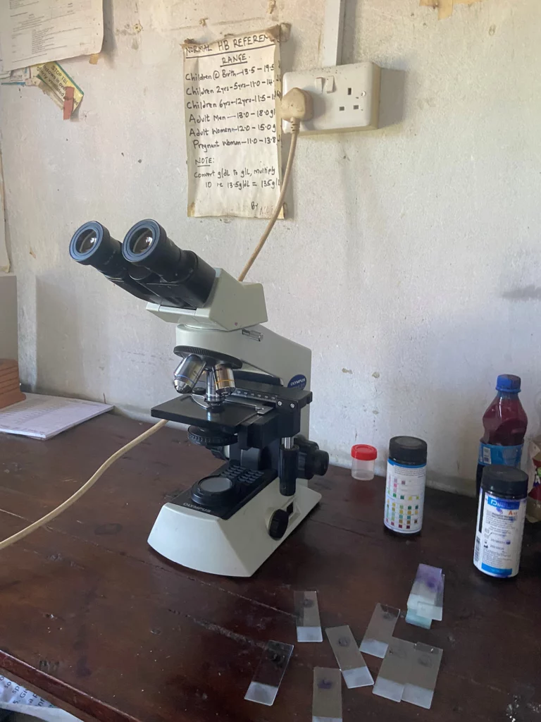 A microscope on a wooden table in a small lab setting, with glass slides, test containers, and a handwritten reference chart for hemoglobin (HB) ranges pinned to the wall above.