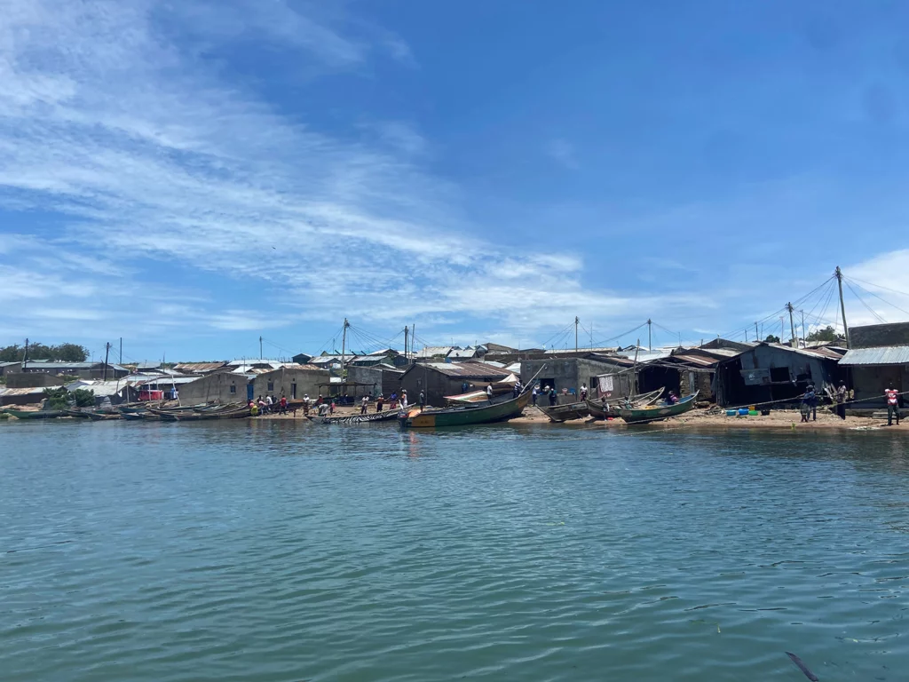 A shoreline view of a fishing village with several wooden boats docked along the water, simple houses with tin roofs, and people gathered near the shore under a bright blue sky.