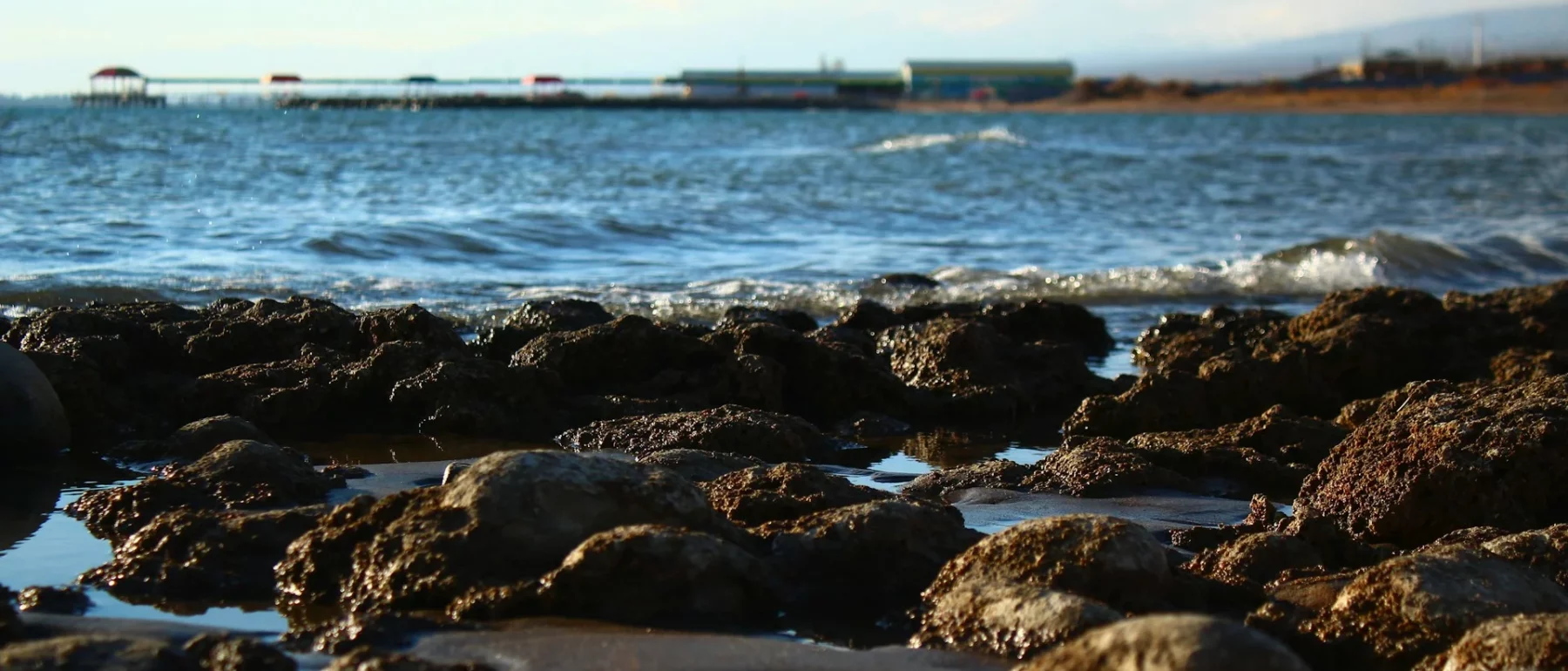 Rocky shoreline with gentle waves and a distant pier over the water.