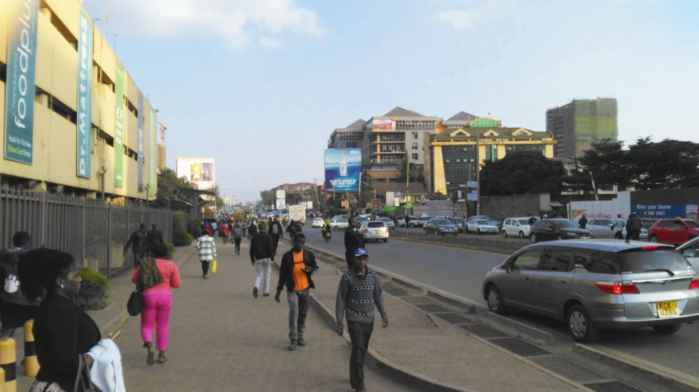 A wide street on a sunny day, with separate wide pavement for pedestrians, and traffic driving away from the camera.