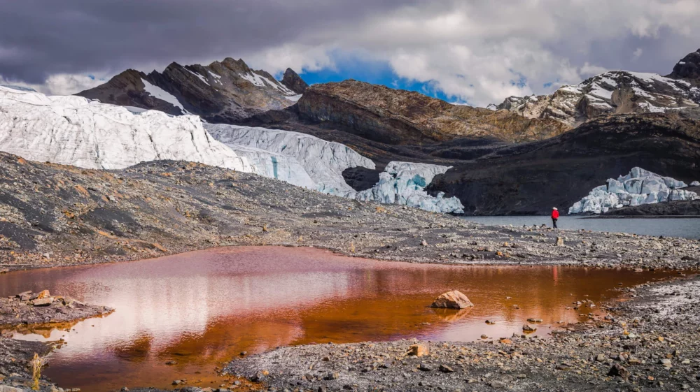 Person in red jacket standing near a glacial lake with reddish water, surrounded by rocky terrain and melting glacier in the Peruvian Andes.