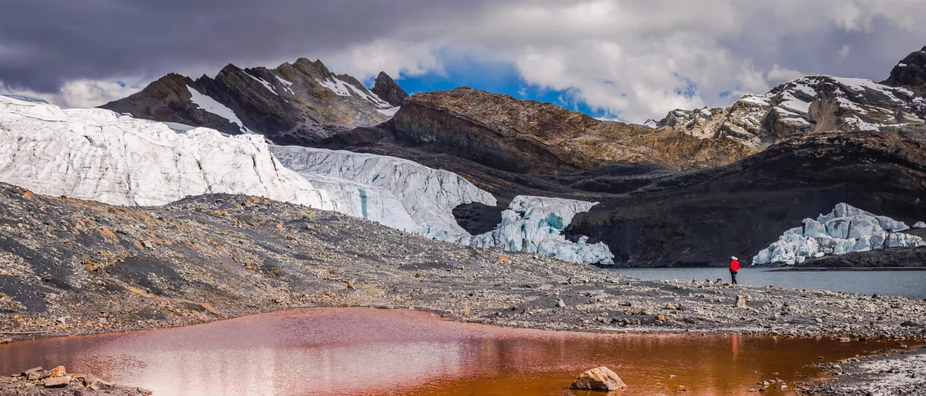 Person in red jacket standing near a glacial lake with reddish water, surrounded by rocky terrain and melting glacier in the Peruvian Andes.