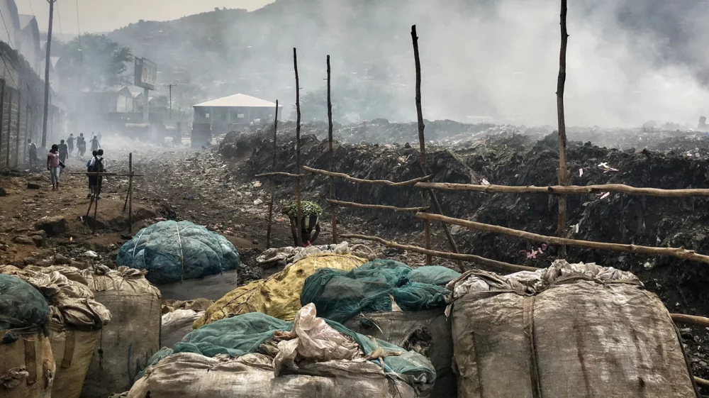 People walking next to a smoking landfill site with rubbish on fire in and large sacks in the foreground in Freetown, Sierra Leone, Africa.