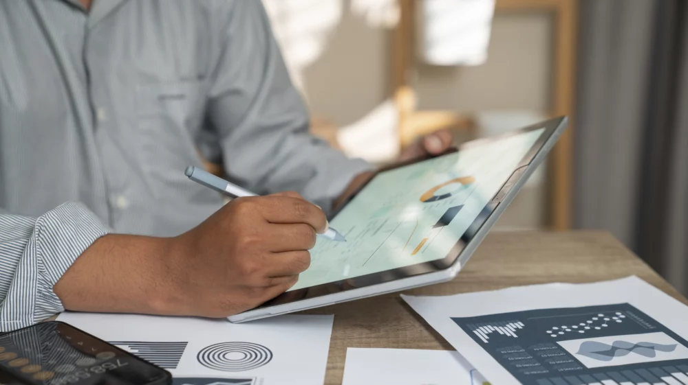 A man working with report charts, analytics. He has a calulator open, he is drawing on a tablet, he has papers with charts in front of him on the table.