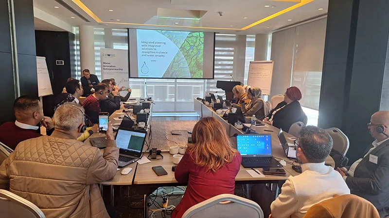 Kim Andersson stands at the front of a conference room presenting to participants seated around a U-shaped table, with a slide on integrated water solutions projected on screen.