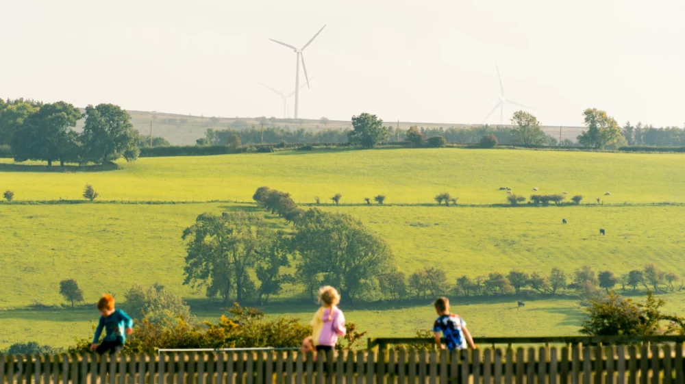 Children in the foreground look out over a green rural landscape with rolling hills, scattered trees, grazing animals, and wind turbines in the distance.