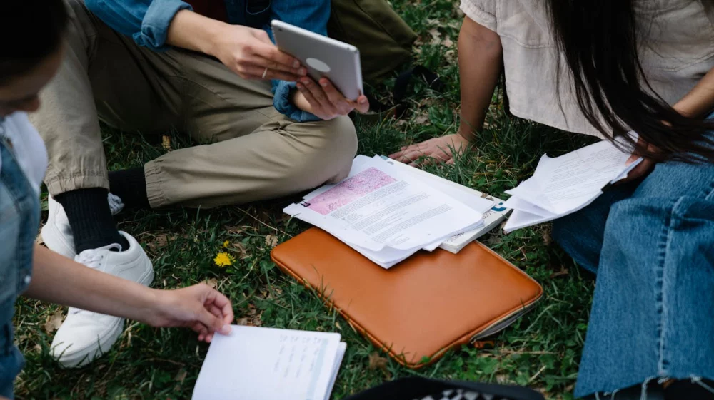 Close up hands of three people sitting on grass with a notebook, tablet, and laptop case.